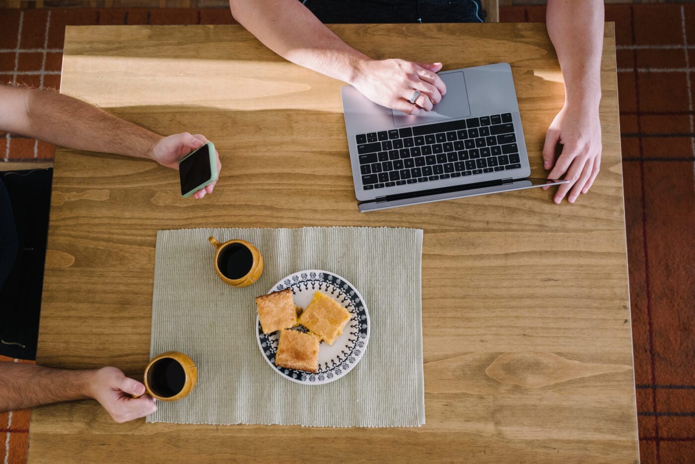 Overhead view of a wooden table with a laptop and trackpad on the right, one person tapping the trackpad while another holds a smartphone on the left, a plate of pastries in the center, and two coffee mugs on a placemat at the bottom left and right.&nbsp;El Blog de Germán Piñeiro